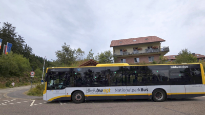 Bwegt Bus gelb, weiss, schwarz bei einer Bushaltestellte mit einem Gebäude im Hintergrund