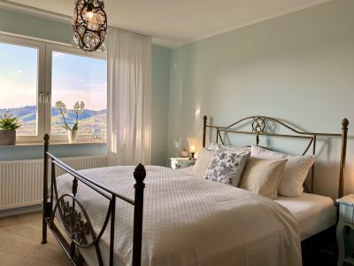 Schlafzimmeransicht, ein Bett mit Decke und Kopfkissen und Ausblick ausm Fenster auf Berge mit Weinreben