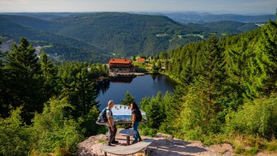 Wanderer an der Augenblick Aussichtstafel mit Blick zum Mummelsee und ins Tal. blauer Mummelsee, Wald, zwei Wanderer und Weitblick, Mummelseehotel ist auch zu sehen.