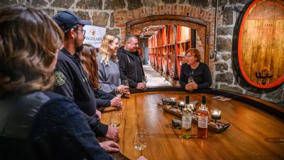 kleine Gruppe mit sechs Personen bei einer Weinprobe in einem Weinkeller, im Hintergrund Weinfässer aus Holz